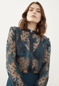 Transparante tealkleurige blouse met bloemborduursels en stippen. Heeft een hoge halslijn, knoopsluiting en lange mouwen.