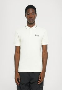 Crème poloshirt met zwarte kraagafwerking en EA7-logo op de linkerkant van de borst, gemaakt van zacht katoen. Klassieke pasvorm met korte mouwen.