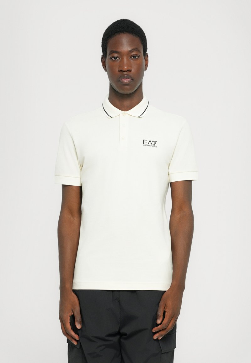 Crème poloshirt met zwarte kraagafwerking en EA7-logo op de linkerkant van de borst, gemaakt van zacht katoen. Klassieke pasvorm met korte mouwen.