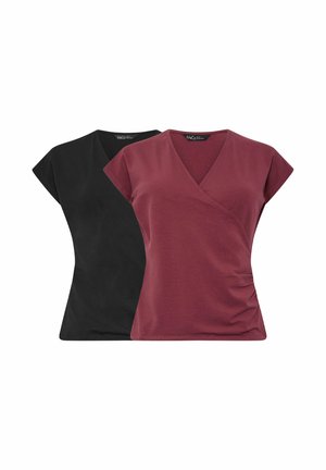 Mujer de pie con una blusa cruzada borgoña de manga corta, vaqueros anchos azules y zapatillas blancas sobre un fondo liso y claro.