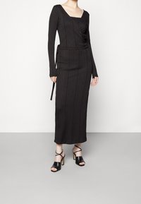 Robe maxi noire à côtes avec des manches longues et un décolleté carré, dotée d'un lien sur le côté. Associée à des sandales à talons ouverts noires.