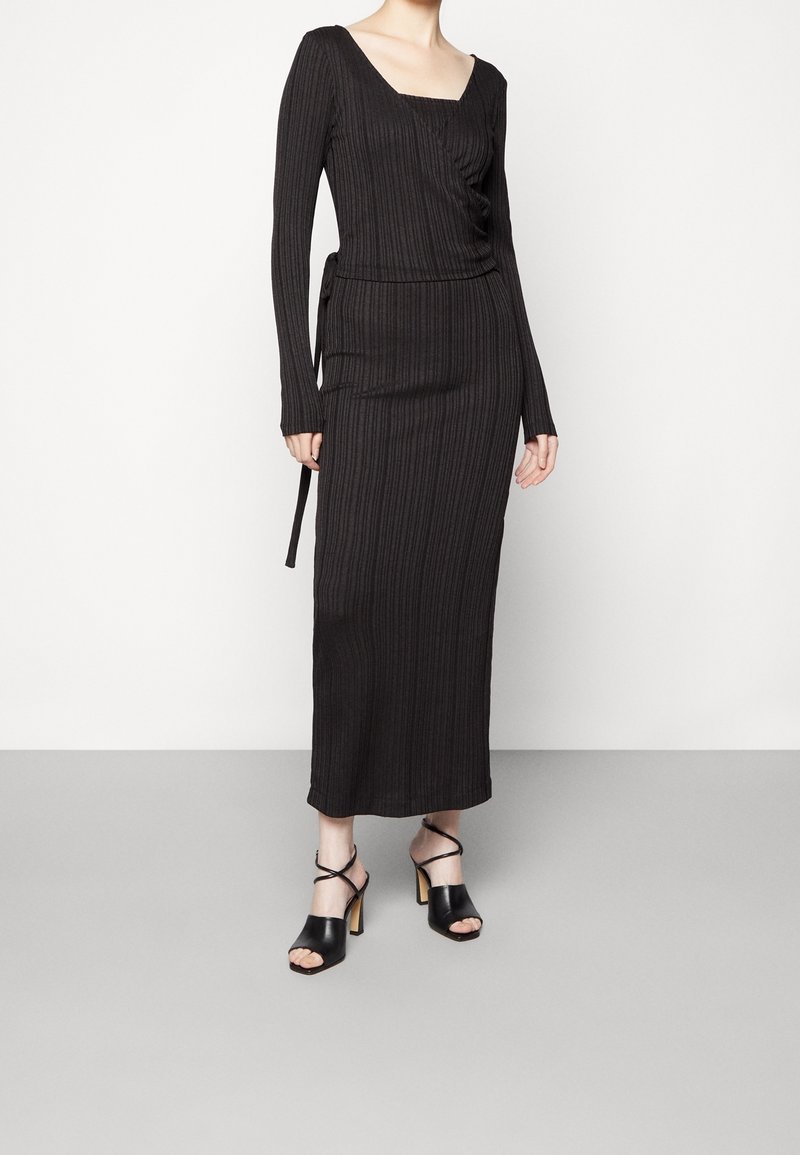 Robe maxi noire à côtes avec des manches longues et un décolleté carré, dotée d'un lien sur le côté. Associée à des sandales à talons ouverts noires.