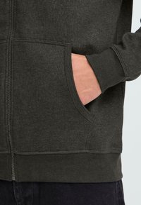 Person's hand partially inside the front pocket of a dark gray zip-up sweatshirt, showing fabric texture and stitching details.