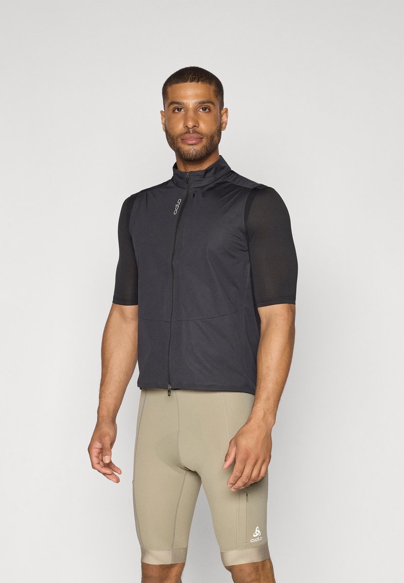 Gilet sans manches noir avec un col montant, fermeture éclair à l'avant et épaules texturées, associé à un short de cyclisme beige avec un logo près de l'ourlet.