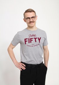 Šedé bavlněné tričko s vínovým potiskem: „Pushování k padesátce je dostatečné cvičení.“ Model nosí černé kalhoty, ruce v bok, mírný úsměv.