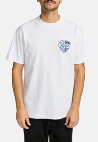 Hvid bomulds t-shirt med et lille blåt og orange grafisk design med palmer og bølger. Rund hals og korte ærmer, afslappet pasform.