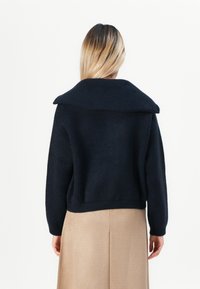Kvinne med langt blondt hår iført en svart genser med bred krage og beige skjørt, vist fra ryggen mot en hvit bakgrunn.