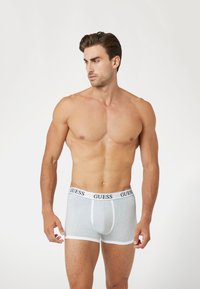Boxer shorts de color gris claro con ribete blanco, con un logo negro "GUESS" en la cinturilla, fabricados en un material suave y elástico.