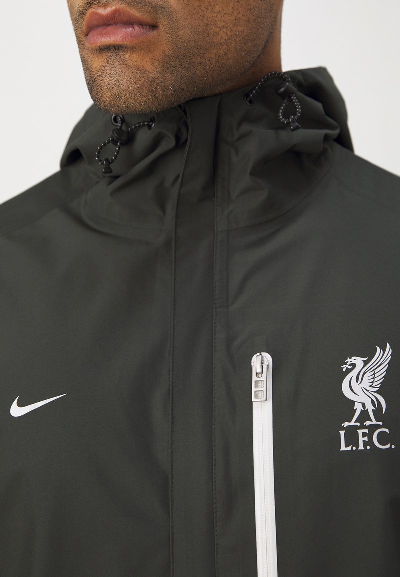 Dunkelgrüne wasserdichte Jacke mit hohem Kragen, weißem Nike-Logo auf der linken Brust und einer weißen Reißverschlusstasche mit einem silbernen Ziehband.