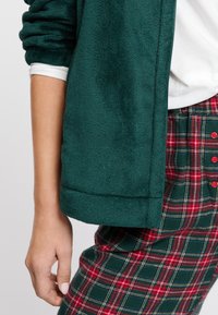Zielona polarowa kurtka z podwiniętymi rękawami i gładką teksturą, w połączeniu z czerwono-zielonymi spodniami w kratę z dużymi guzikami i wzorem w kratkę.