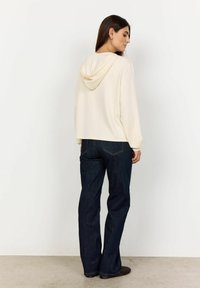 Felpa color crema con cappuccio e cordoncino, indossata con jeans blu scuri. La felpa ha una vestibilità comoda e una trama morbida.