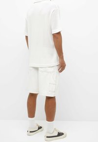 Polo blanca de malla con mangas cortas, combinada con pantalones cortos cargo blancos hasta la rodilla que cuentan con bolsillos laterales. Calzado casual completa el atuendo.