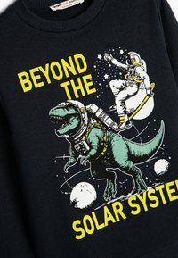 Sudadera de color navy con un gráfico de un dinosaurio en un traje de astronauta y el texto " MÁS ALLÁ DEL SISTEMA SOLAR" en amarillo brillante.