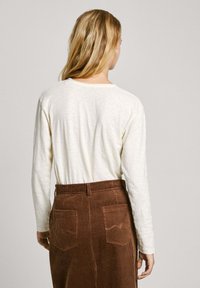 Off-white ilgo rankovėmis marškiniai su apvaliu kaklu, derinami su rudais šortais iš šviesaus veliūro, turinčiais vertikalius briaunų raštus ir dvi nugarines kišenes.