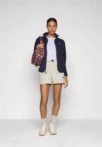 Giacca in pile blu navy, camicia bianca, pantaloni corti beige chiaro, sneakers color crema, che tiene una borsa a tracolla bordeaux. Design semplice, caratteristiche funzionali e tasche.