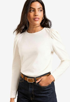 Cremfarbener Langarmoberteil mit gepufften Schultern, kombiniert mit dunklen Denim-Jeans und einem braunen Gürtel. Glattes Material mit einem taillierten Design.