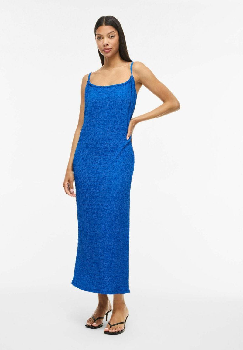 Femme en robe maxi bleu roi texturée sans manches à fines bretelles, debout avec une main sur la hanche et portant des sandales noires à talons.