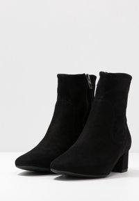 Stivali alla caviglia in suede nera con tacco a blocco e cerniere laterali, esposti su una superficie bianca su uno sfondo uniforme.