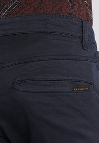 Tumman siniset housut, joissa on sileä tekstuuri, varustettu vetoketjulla takataskussa ja pienellä ruskealla merkkilapulla vyötäröllä.