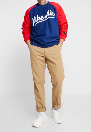 Persona che indossa una felpa Nike Air blu e rossa, pantaloni beige risvoltati e sneakers bianche, in piedi con le mani nelle tasche su uno sfondo grigio.