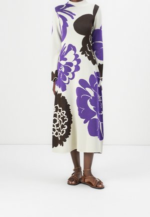 Long white dress with large purple and dark brown floral patterns, long sleeves, paired with brown ankle-strap sandals on dark skin.