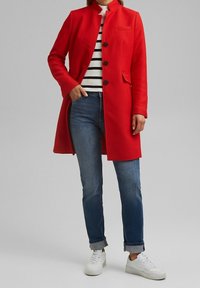 Rode mantel van een wolmix met hoge kraag, enkele knopen aan de voorkant en zijzakken, gecombineerd met een gestreepte trui en blauwe jeans.