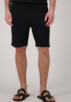 Zwarte, gestructureerde shorts met een elastische tailleband en trekkoord, voorzien van zijzakken en een rechte snit. Draagt bij zwarte sandalen.
