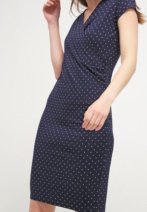 Femme portant une robe bleu marine à pois blancs, longueur genou, manches courtes, et un corsage style portefeuille, debout devant un fond clair uni.