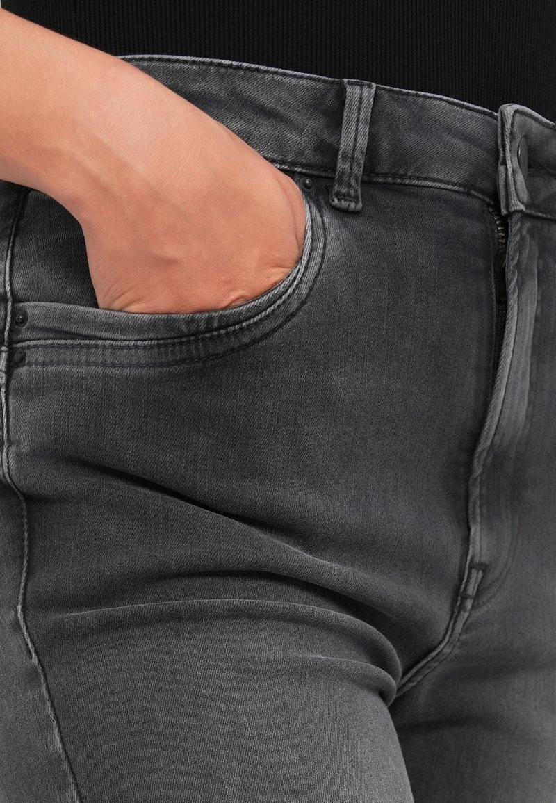 Graue Denim-Jeans mit einer Nahaufnahme der Tasche und einer Hand, die darin ruht. Die Jeans zeigen einen texturierten Stoff und sichtbare Nähte.