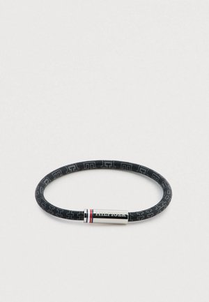 Pulsera de goma negra con patrón de logo en relieve, terminal metálico plateado con franjas de acento rojas y blancas. Forma circular.