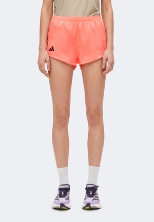 Person trägt leuchtend korallenfarbene Sportshorts, weiße Crew-Socken und lila Laufschuhe und steht vor einem schlichten Hintergrund.