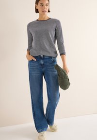 Grauer Strickpullover, weit geschnittene blaue Jeans und beige Sneakers. Hält eine grüne, gesteppte Tasche. Lässiges Outfit mit entspannter Passform.