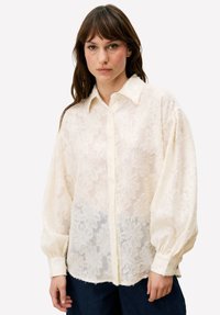 Crèmekleurige blouse met een gestructureerd kanten patroon, knoopsluiting aan de voorkant en oversized mouwen. Zachte, doorschijnende stof met een licht glanzende afwerking.
