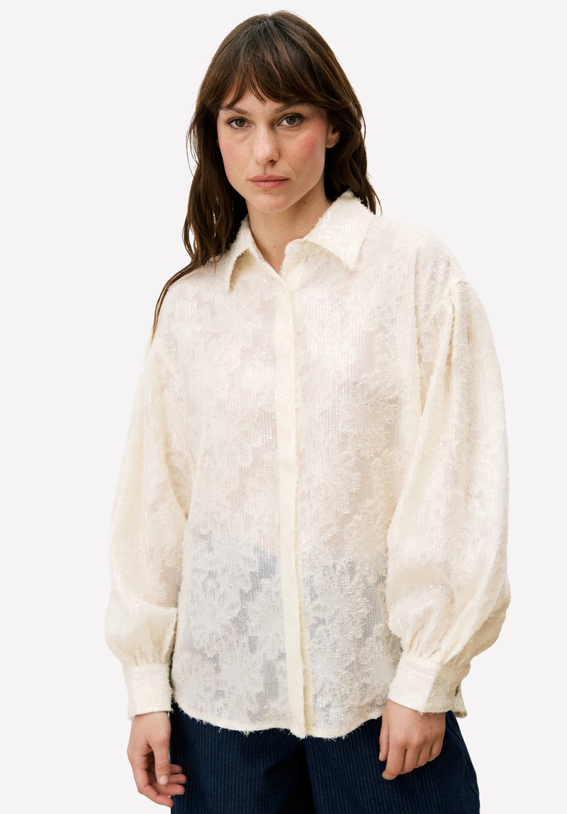 Crèmekleurige blouse met een gestructureerd kanten patroon, knoopsluiting aan de voorkant en oversized mouwen. Zachte, doorschijnende stof met een licht glanzende afwerking.