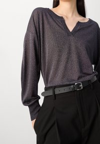 Frau in einem dunkel schimmernden Langarmoberteil mit einem Schlitz-Ausschnitt, das in hochgeschnittene schwarze Hose mit einem schwarzen Gürtel gesteckt ist.