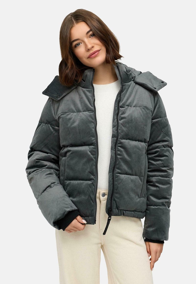 Grijze pufferjack met een gequilt ontwerp en een capuchon. Beschikt over een voorzijde rits en zwarte manchetten. G gedragen over een lichte top en beige broek.