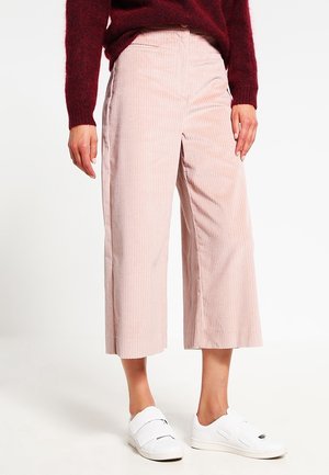 Persoon die lichtroze corduroy broek met wijde pijpen en cropped lengte draagt, een bordeauxrode trui en witte sneakers met klittenbandsluiting, staand op een witte achtergrond.