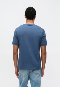 Modra bombažna majica z okroglim izrezom in kratkimi rokavi. Gladek tekstura, pogled od zadaj. Kombinirana z svetlo modrimi bledenimi kavbojkami.