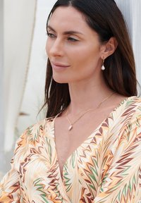 Gold chain necklace with a pearl pendant, matching pearl drop earrings. Model wears a patterned, v-neck blouse in orange, green, and white.