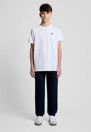 Teenagedreng står med fronten mod kameraet iført en hvid T-shirt med gul logo, mørke bukser og hvide sneakers mod en ensfarvet grå baggrund.