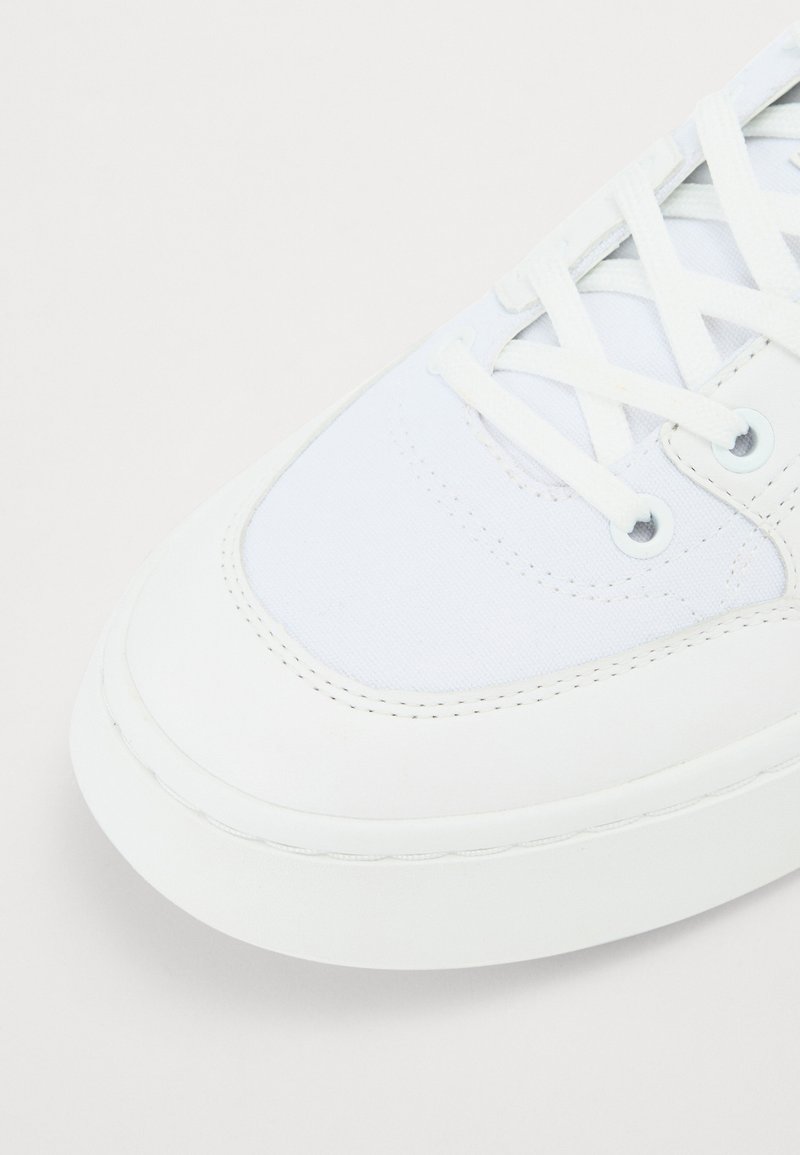 Sneaker blanche avec un dessus en cuir lisse et en toile, bout rond, lacets blancs plats et détails de couture subtils sur une semelle en caoutchouc élégante.