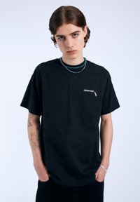 Camiseta de algodón negra de manga corta, con un pequeño texto gráfico blanco en el pecho y un corte relajado. Lleva puesta una serie de collares de cuentas en capas.