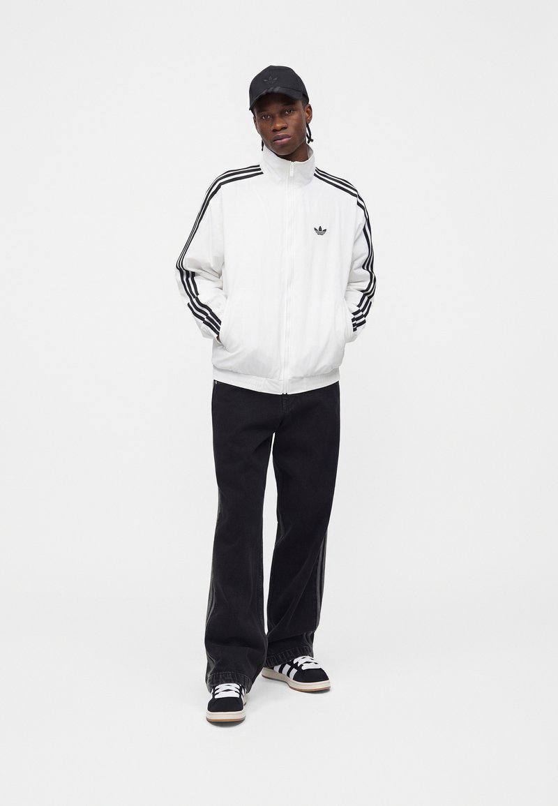 Ung mand iført en hvid Adidas trackjakke med sorte striber, sorte bukser, sorte Adidas sneakers og en sort kasket, stående med hænderne i lommerne.