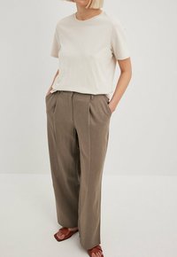 Jasno beżowa koszulka w połączeniu z luźnymi, brązowymi spodniami z wysokim stanem, które mają przednie zakładki i boczne kieszenie. Całość dopełniają brązowe płaskie sandały.