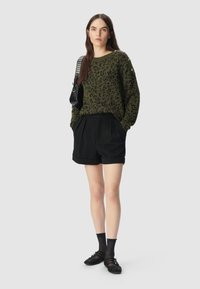Vrouw in een olijfgroene trui met luipaardprint, zwarte geplooide short, zwarte doorschijnende sokken, zwarte schoenen en een zwarte schoudertas met zilveren studs.