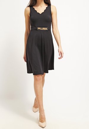 Femme portant une robe noire sans manches longueur genoux avec un décolleté festonné et une ceinture à boucle rectangulaire, associée à des escarpins beige.