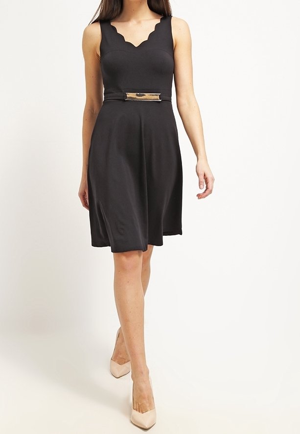 Femme portant une robe noire sans manches longueur genoux avec un décolleté festonné et une ceinture à boucle rectangulaire, associée à des escarpins beige.