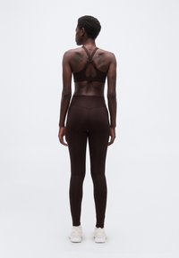 Γυναίκα με καστανό σκούρο αθλητικό σουτιέν με σταυρωτούς ιμάντες, συνδυαστικό κολάν και λευκά αθλητικά παπούτσια, σε ουδέτερο φόντο, με την πλάτη να κοιτάει.