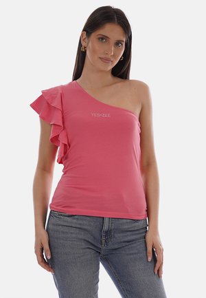 Donna con top rosa monospalla con dettaglio a balze sul lato sinistro e jeans grigi, in piedi contro uno sfondo semplice.