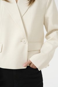 Beige pielāgota žakete ar iedvesmotiem klapjiem, vienu pogas aizdari un diviem sānu kabatām, saskaņota ar melniem biksēm. Gluda auduma tekstūra.